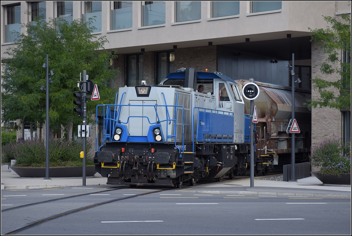 Gravita Am 847 003 'Littau' schiebt einen schweren Stahlzug von der Emmenwaid durch das Eckhaus in den Bahnhof Emmenbrücke. Das kantige Design der Gravita passt zum modernen Eckhaus. August 2021.
