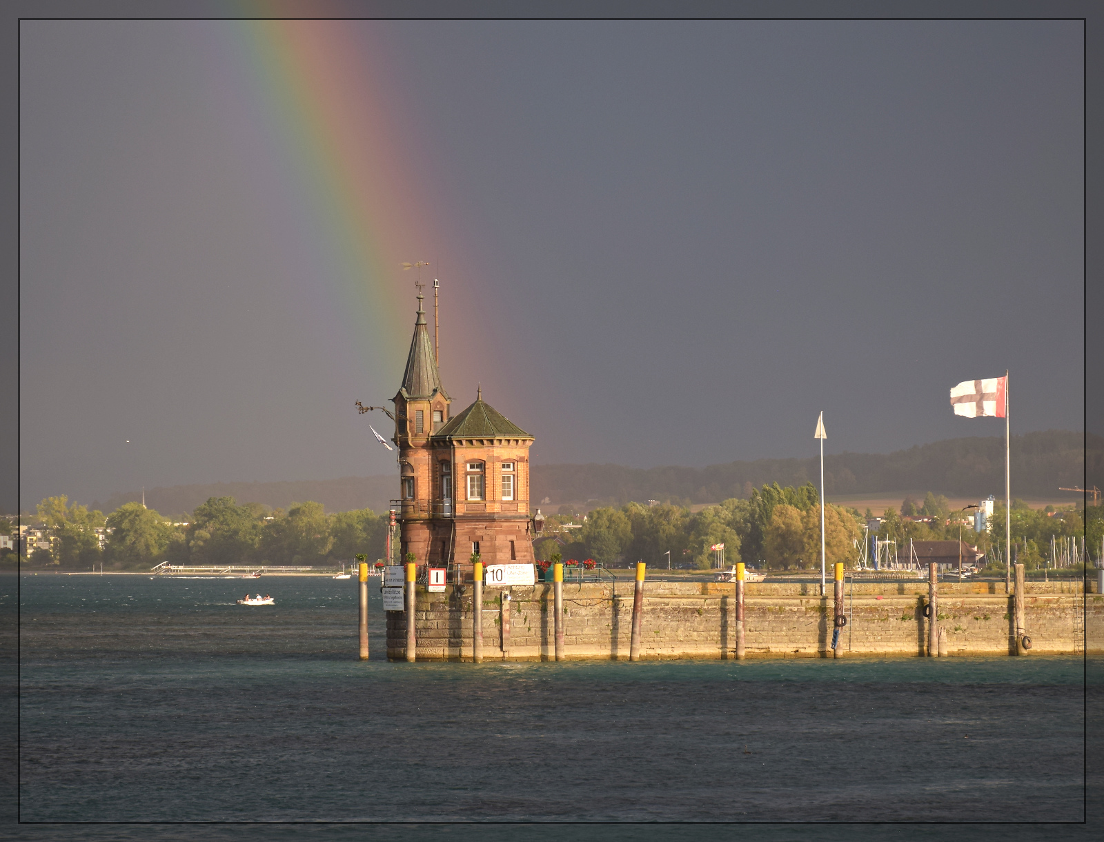 Die Sonne lugt bereits unter einem einem aufkommenden Gewitter hindurch und beleuchtet die Hafeneinfahrt von Konstanz. Dahinter in schönsten Farben, vom Sandsturm gedämpft, ein Regenbogen. August 2026.