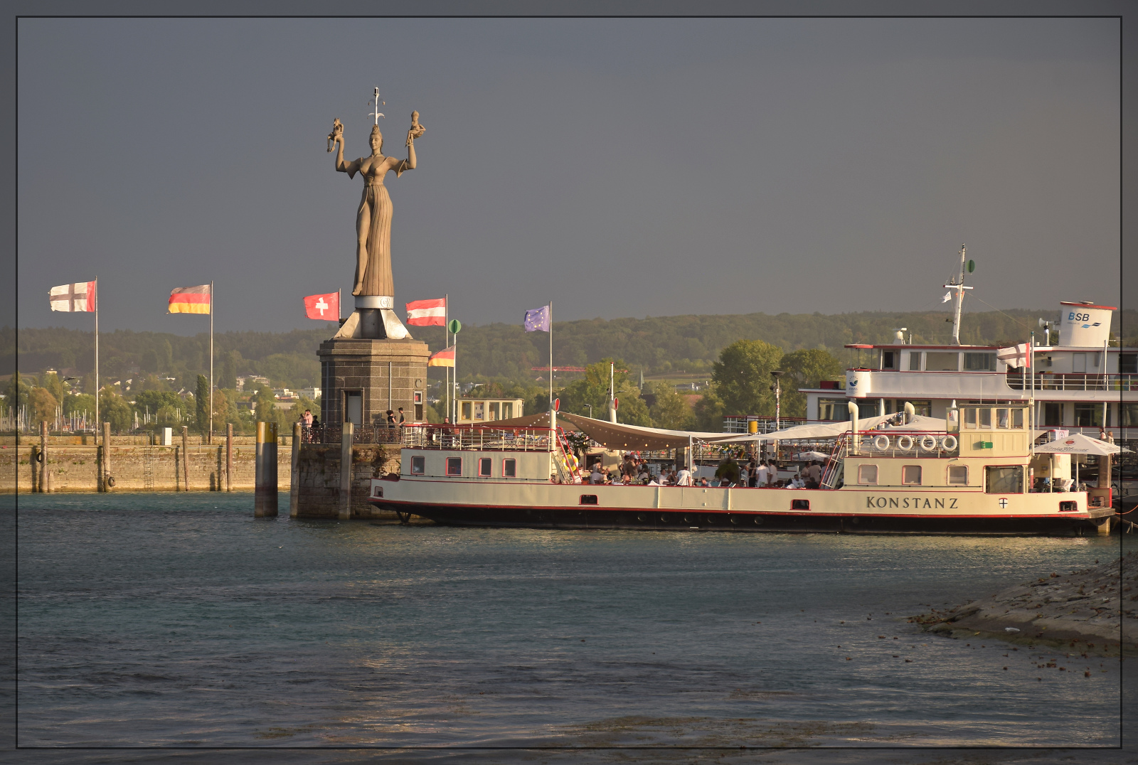 Die Sonne lugt bereits unter einem einem aufkommenden Gewitter hindurch und beleuchtet die Hafeneinfahrt von Konstanz, während der Sturm den ganzen Staub aus dem Stadtgarten auf den See treibt. August 2026.