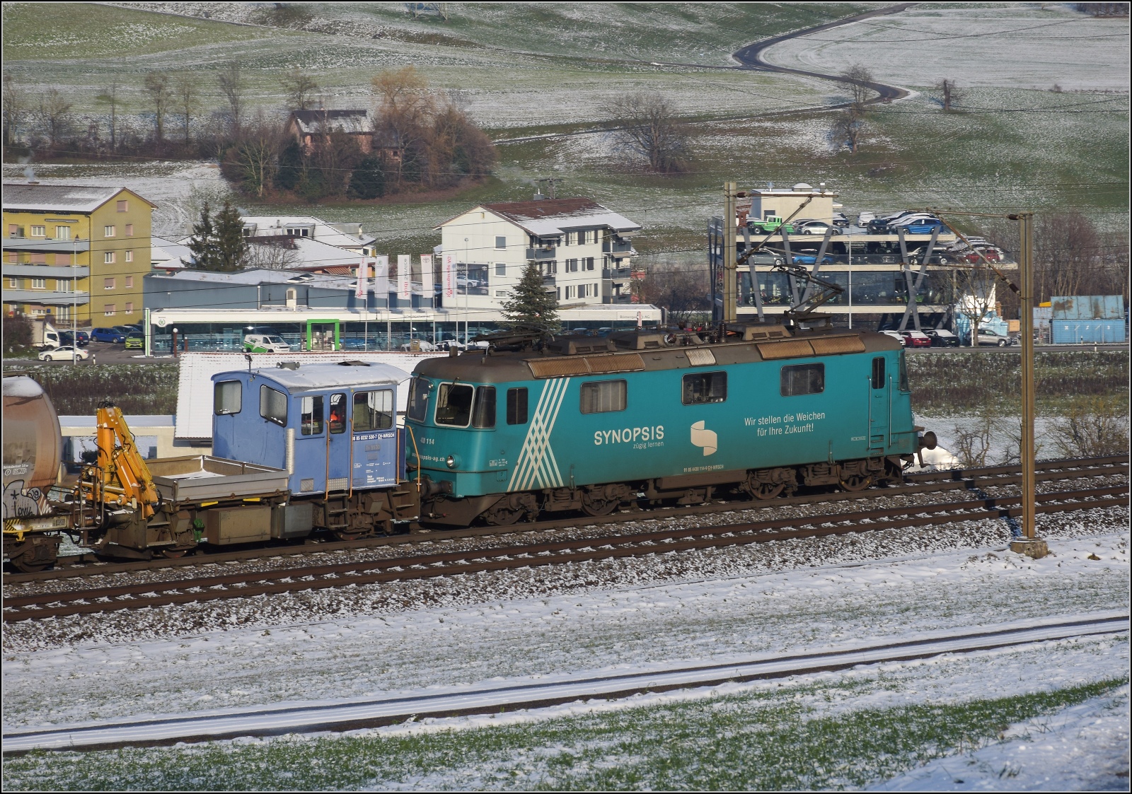 Re 430 115 von WRS mit Synopsis-Werbung schleppt die Rangierhilfe Tm III 9530 mit über den Bözberg. Frick, Dezember 2022.