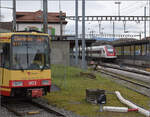 Die Strassenbahn  Be 4/8 003  alias 94 800 450 003-9 D-TVYS wartet in Chavornay auf die Fahrgäste der S-Bahnen während auf der Jurasüdfusslinie noch die ICN RABDe 500 015 'Jean-Jacques Rousseau' und RABDe 500 029 'Eduard Spelterini' vorbeiziehen. Dezember 2025.